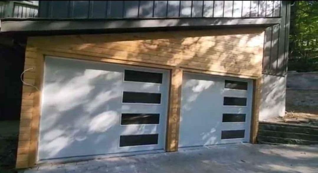 Modern two-car garage with wood accents and horizontal glass panels, surrounded by greenery.