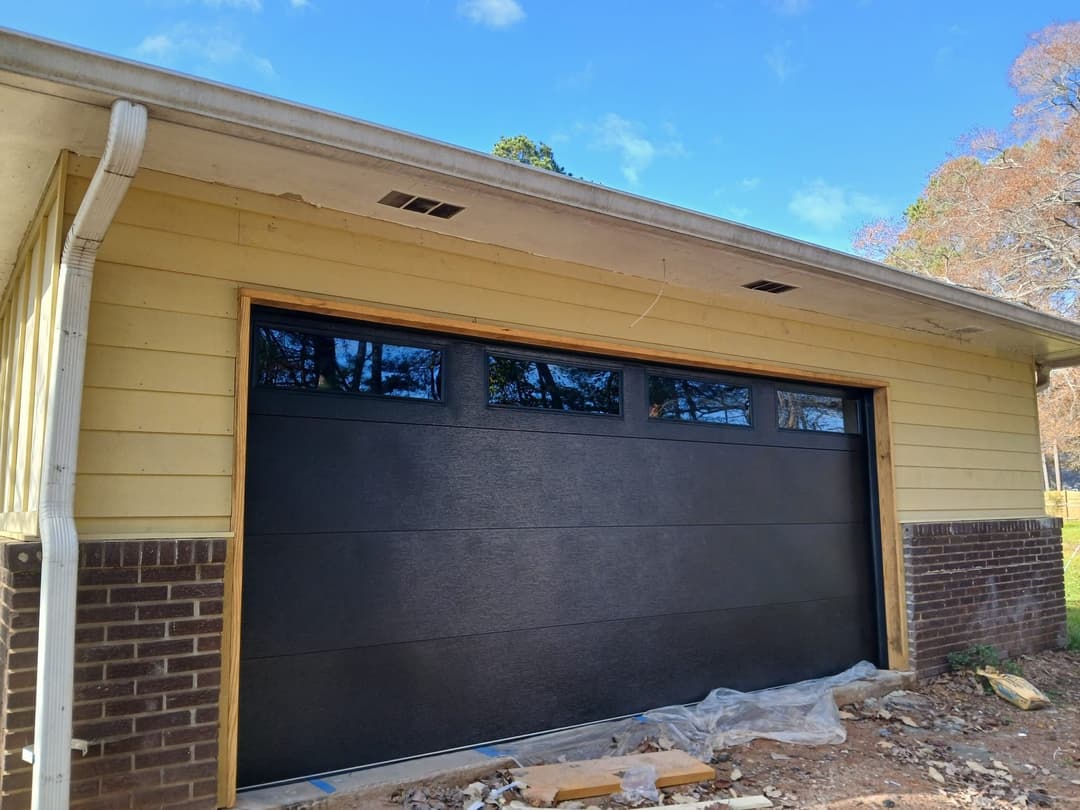 Modern black garage door installation on a yellow house with brick accents, under blue sky.