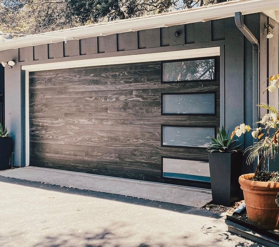 Modern garage door with wooden texture, glass panels, and stylish planters outside.