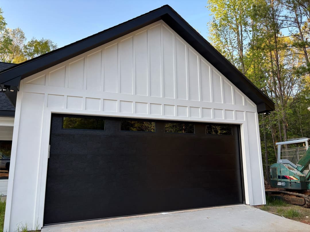 Modern black garage door on a white house with a sloped roof and forest backdrop.