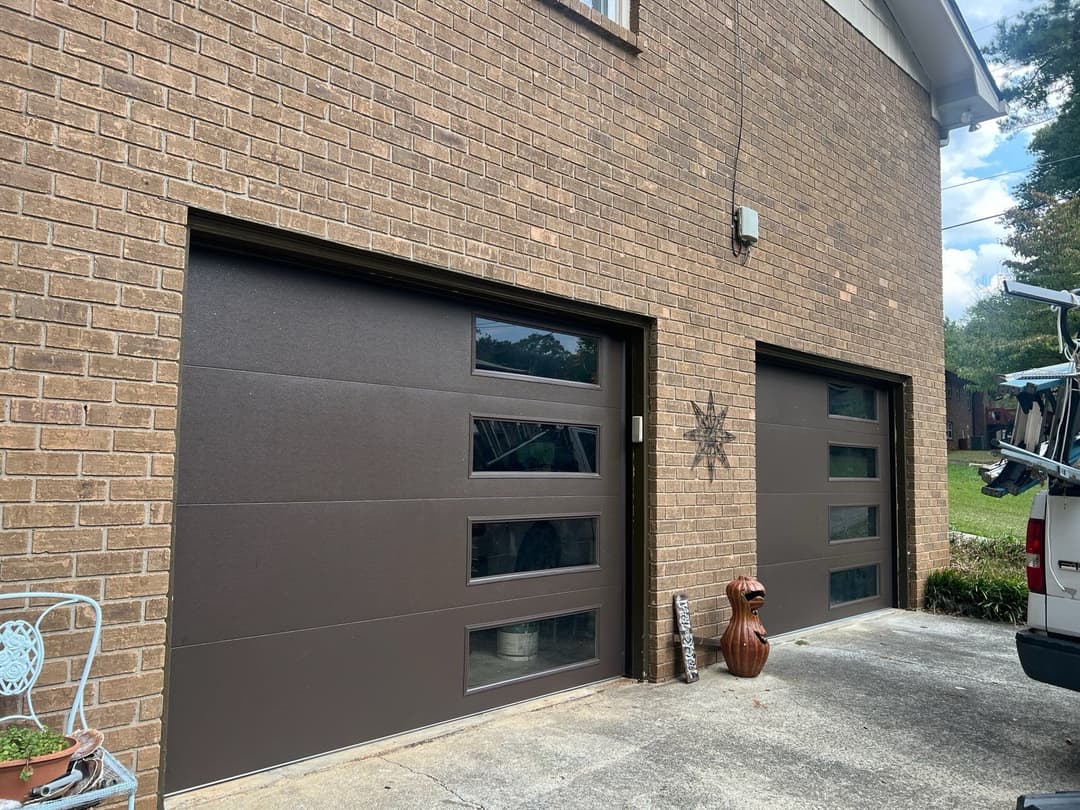 Modern brown garage doors with windows on a brick house exterior. Green lawn in the background.