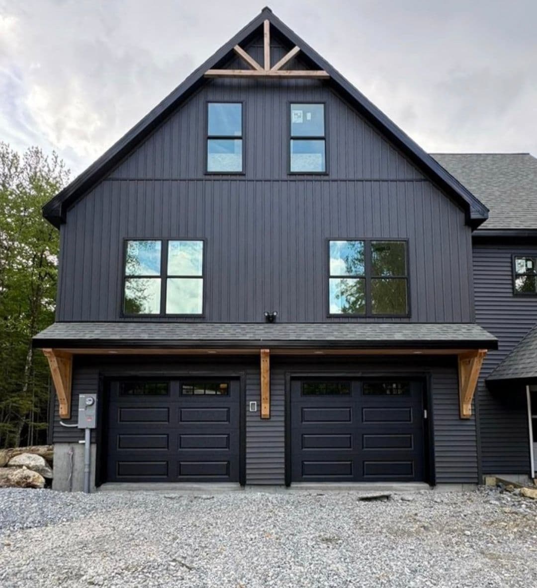 Modern black wooden house with large windows and double garage doors, surrounded by greenery.