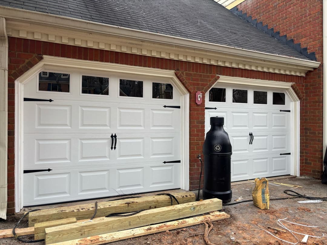 White garage doors with decorative black handles, bricks, and construction materials nearby.