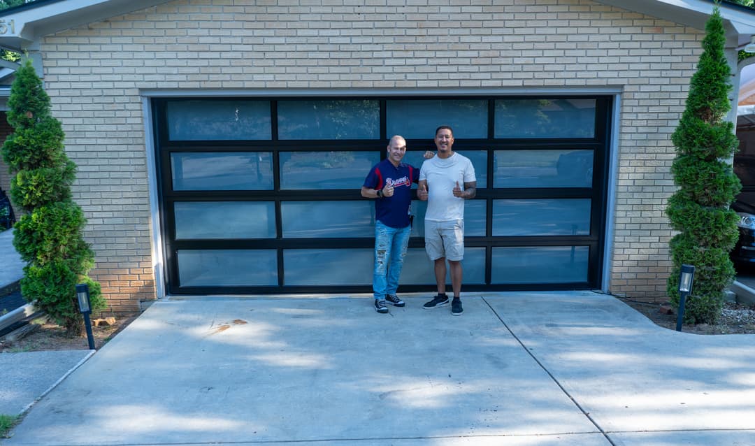 Two men standing in front of a modern garage door, smiling and holding drinks.