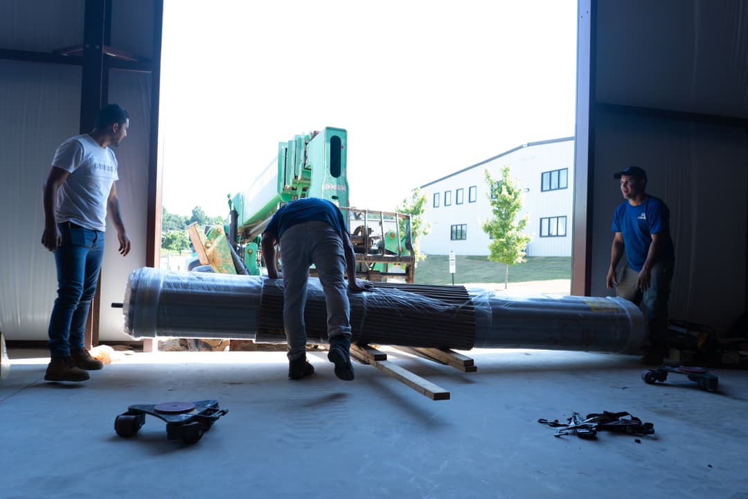 Workers unloading rolled materials in a warehouse with construction equipment in the background.