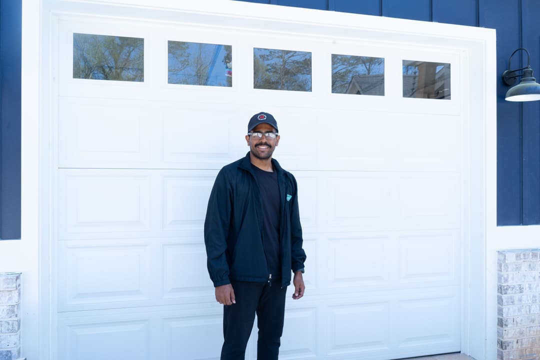 Man standing in front of a modern home garage door, wearing a black jacket and cap.