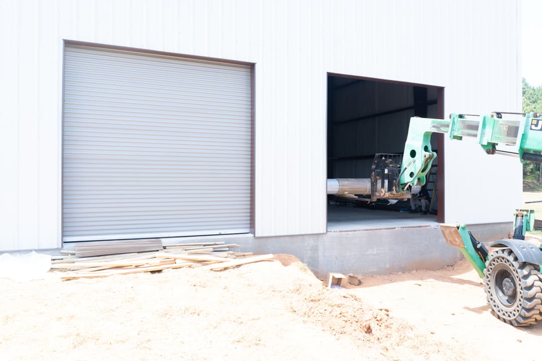 Forklift unloading materials from a warehouse with roll-up doors on a construction site.