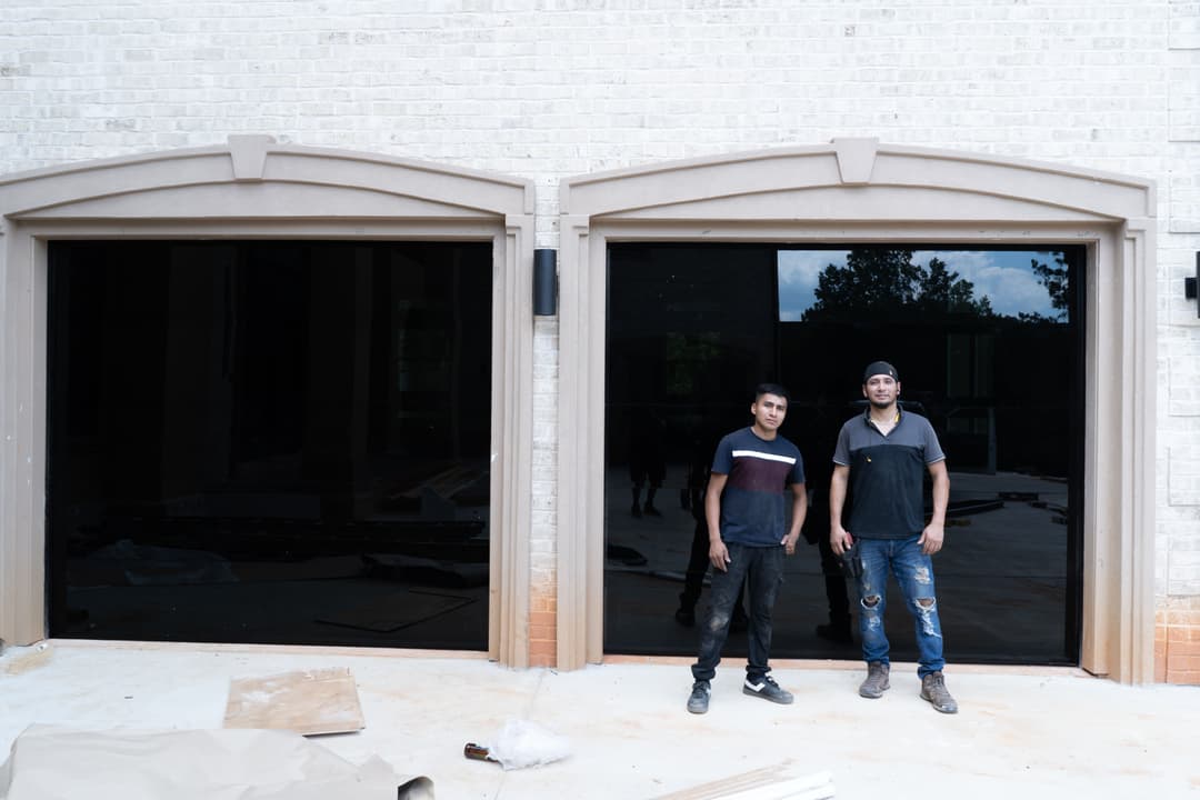Two construction workers stand in front of large, dark window frames at a building site.