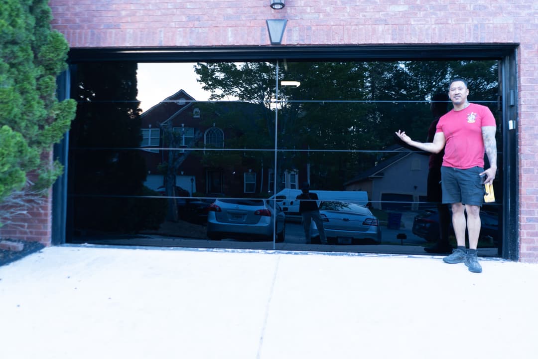 Man gesturing near a modern garage door with reflective glass panels and parked cars in view.