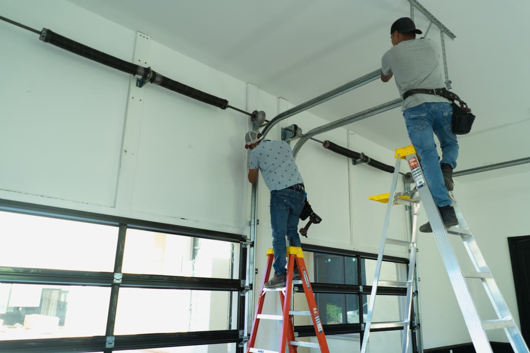 Two workers using ladders to install garage door hardware in a modern garage.