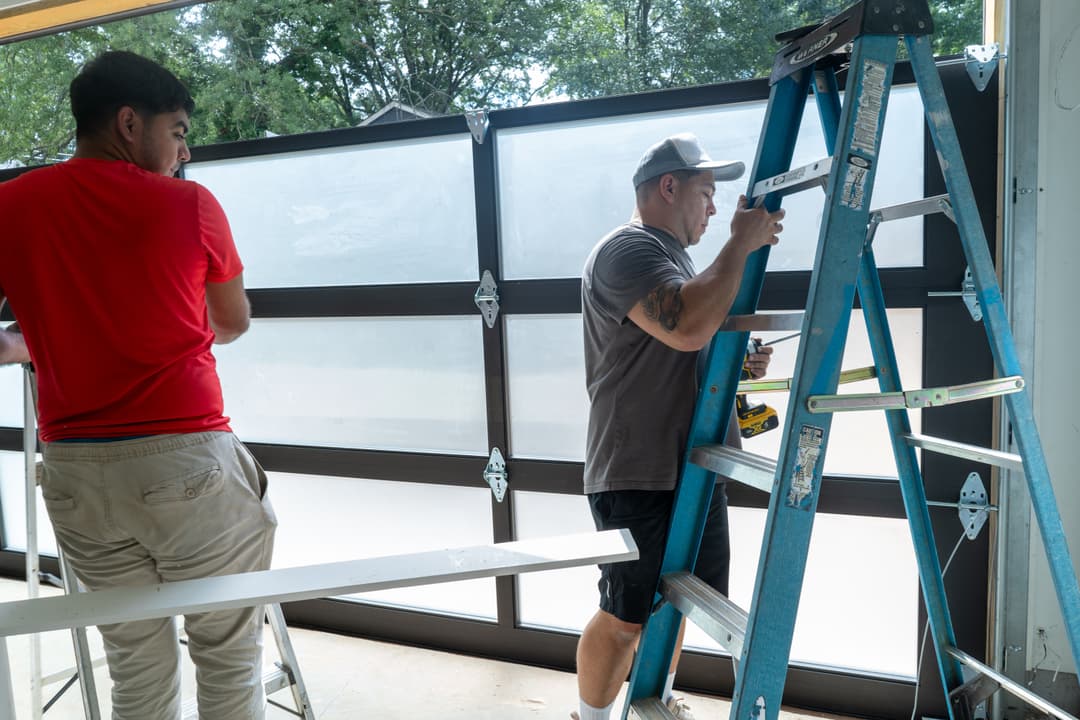 Workers installing a large garage door, one on a ladder with tools, the other assisting nearby.