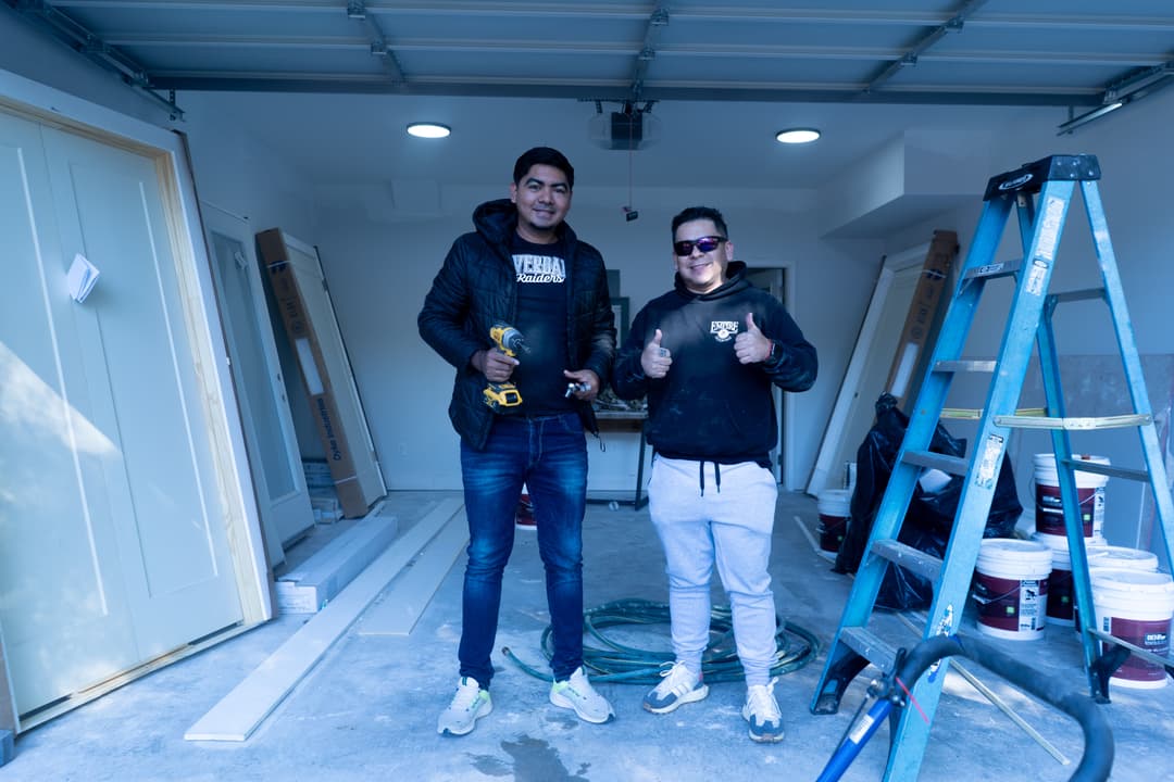 Two men smiling and giving thumbs up in a garage renovation, tools and materials visible.