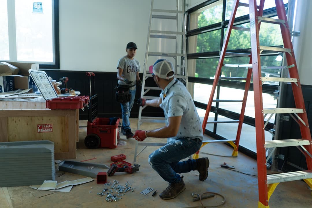 Construction workers on-site, one kneeling with tools, another standing with a ladder.
