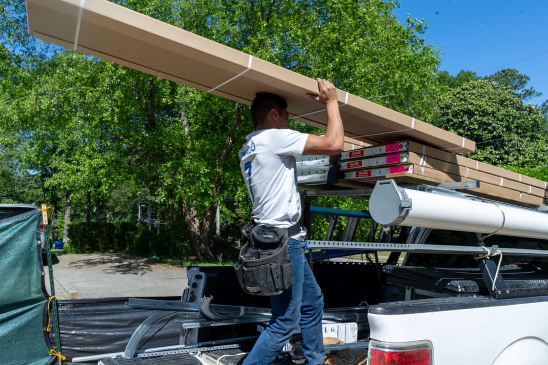 Man lifting long, packaged materials onto a truck bed in a sunny outdoor setting.