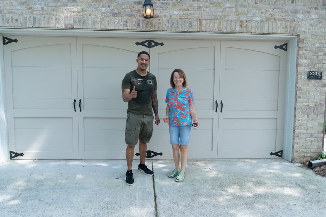 Man and woman smiling in front of a garage, thumbs up, bright outdoor setting.