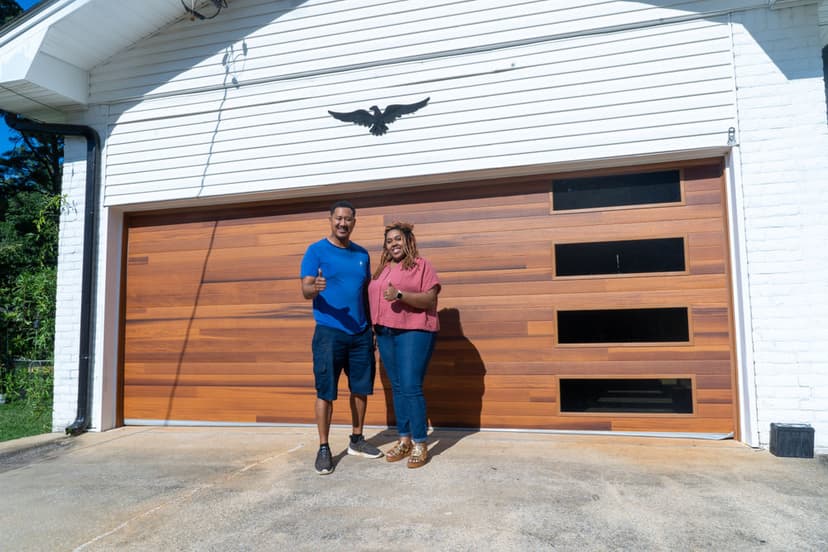 Two people giving thumbs up in front of a modern wooden garage door and white house.